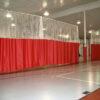 Indoor gym with red privacy curtains forming a partition and white netting overhead several lights above it
