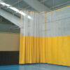 Yellow and transparent fabric curtain divider hangs from the ceiling in a spacious gym, separating workout areas on a blue floor.
