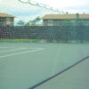 Outdoor tennis court with green mesh fence and netting, buildings in the background.