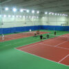 Indoor tennis facility with red courts, green surround, blue divider curtains, and players on two courts under bright overhead lights.