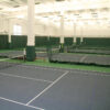 Indoor tennis facility with multiple courts separated by nets and green partitions, bright ceiling lights.