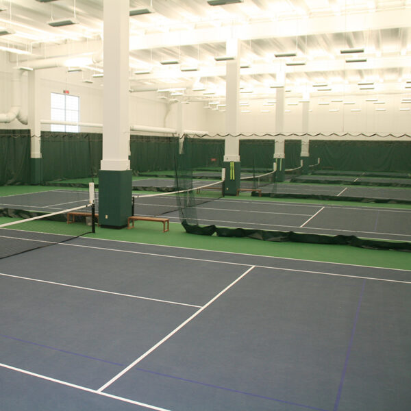 Indoor tennis facility with multiple courts separated by nets and green partitions, bright ceiling lights.