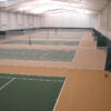Indoor tennis facility with multiple green courts separated by nets and a brown surrounding floor under a bright, white ceiling.