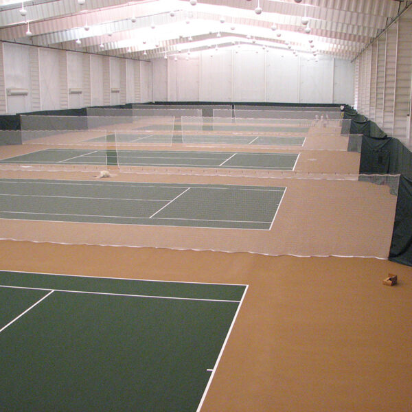 Indoor tennis facility with multiple green courts separated by nets and a brown surrounding floor under a bright, white ceiling.