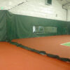 Indoor batting cage inside a gym: green padded walls, black netting overhead, and a red floor. A windowed passage shows a logo in the background.