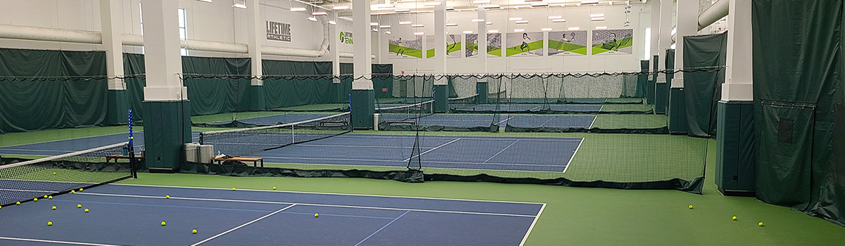 Indoor tennis facility with blue courts, nets, and scattered tennis balls on a green floor.